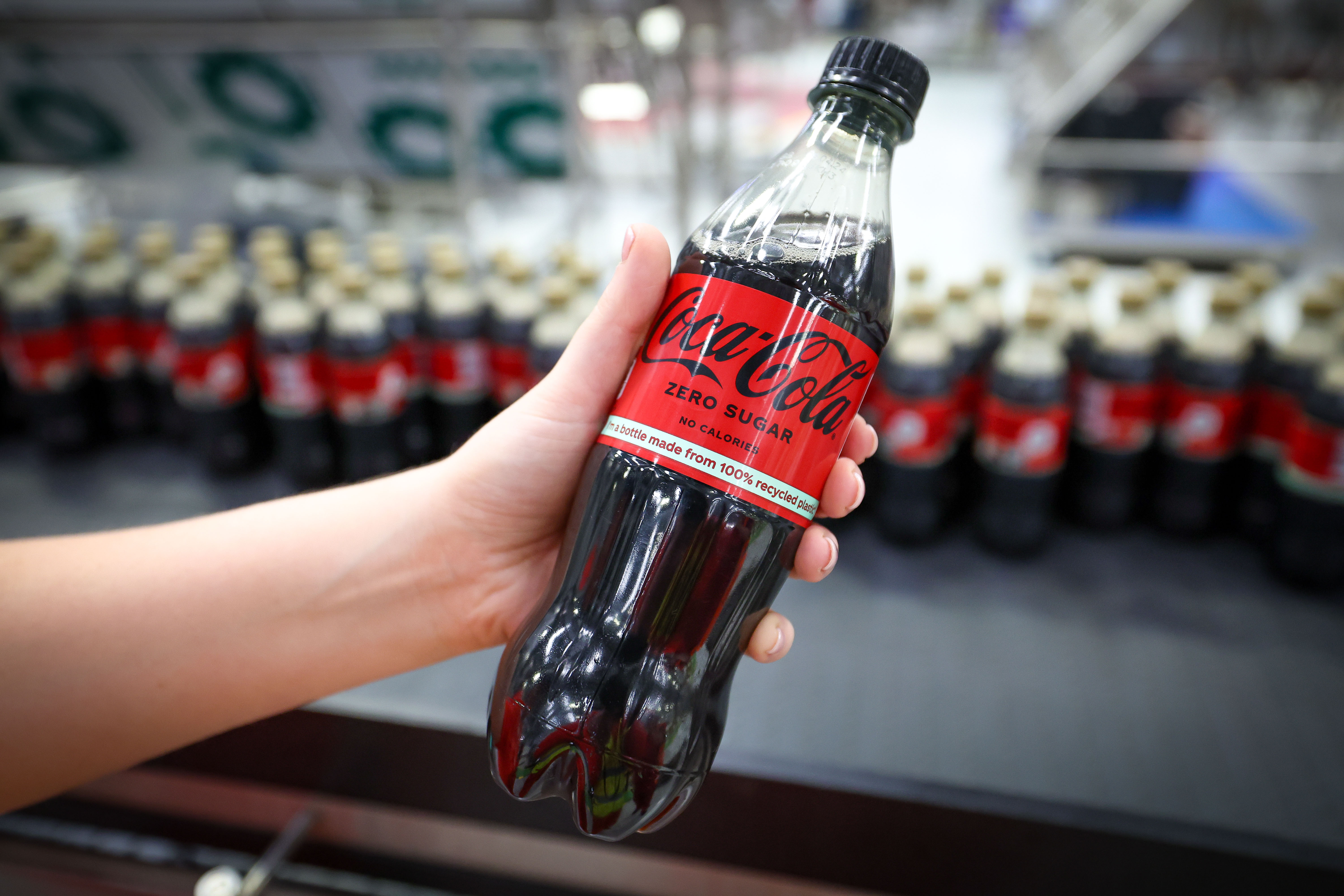 Close-up of a hand holding a Coke Zero Sugar bottle with the label "I'm a bottle made from 100% recycled plastic* (excluding cap and label) with several other bottles blurred in the background on a production line.